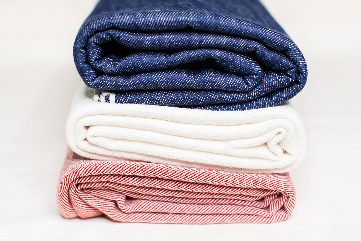 A stack of three folded fabrics of different weights, patterns and colors - double slub selvedge denim on top.