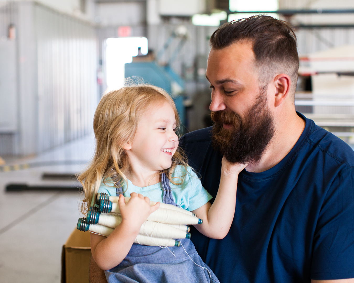 color-photography-craftsman-holding-smiling-young-blonde-daughter-who-is-holding-shuttle-bobbins-while-also-playing-with-his-beard