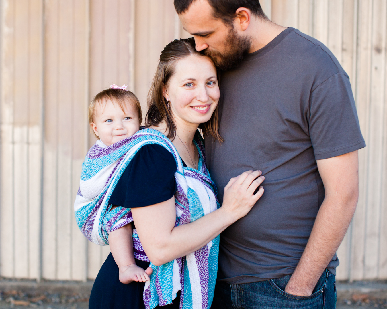 photograph-smiling-young-woman-wearing-colorful-baby-carrier-tall-man-kissing-her-forehead