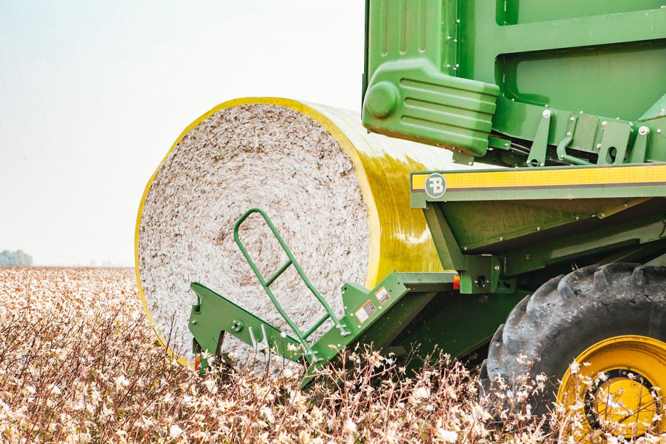 photograph of a cotton field with a green and white tractor transporting a full bale of cotton