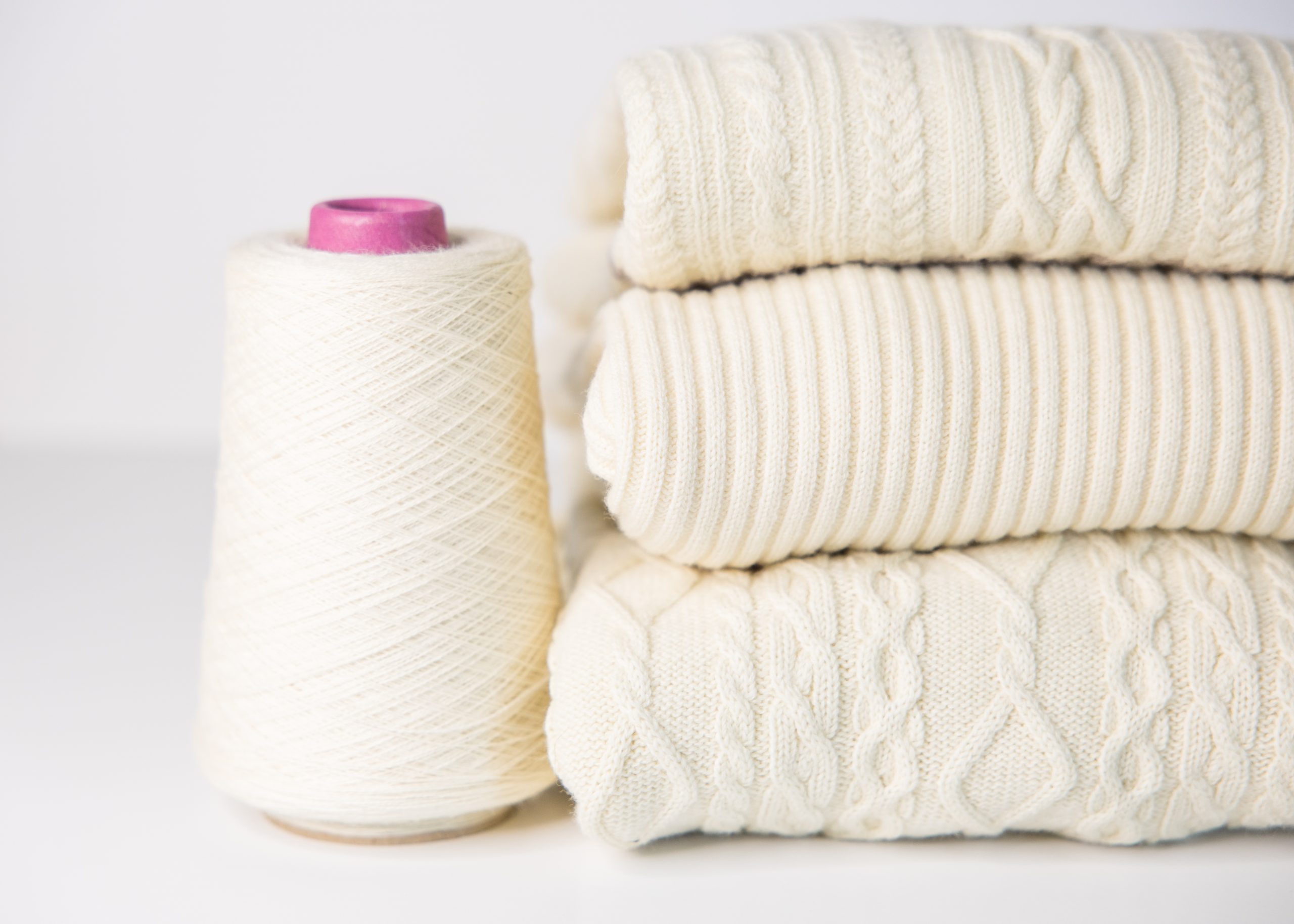 Detailed photograph of oyster white wool, Multi-cable fine knit Wholegarment three sweaters folded and stacked next to a spool of yarn